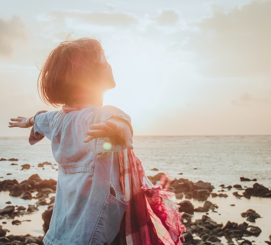 Woman with arms outstretched facing the ocean at sunset on a rocky shore