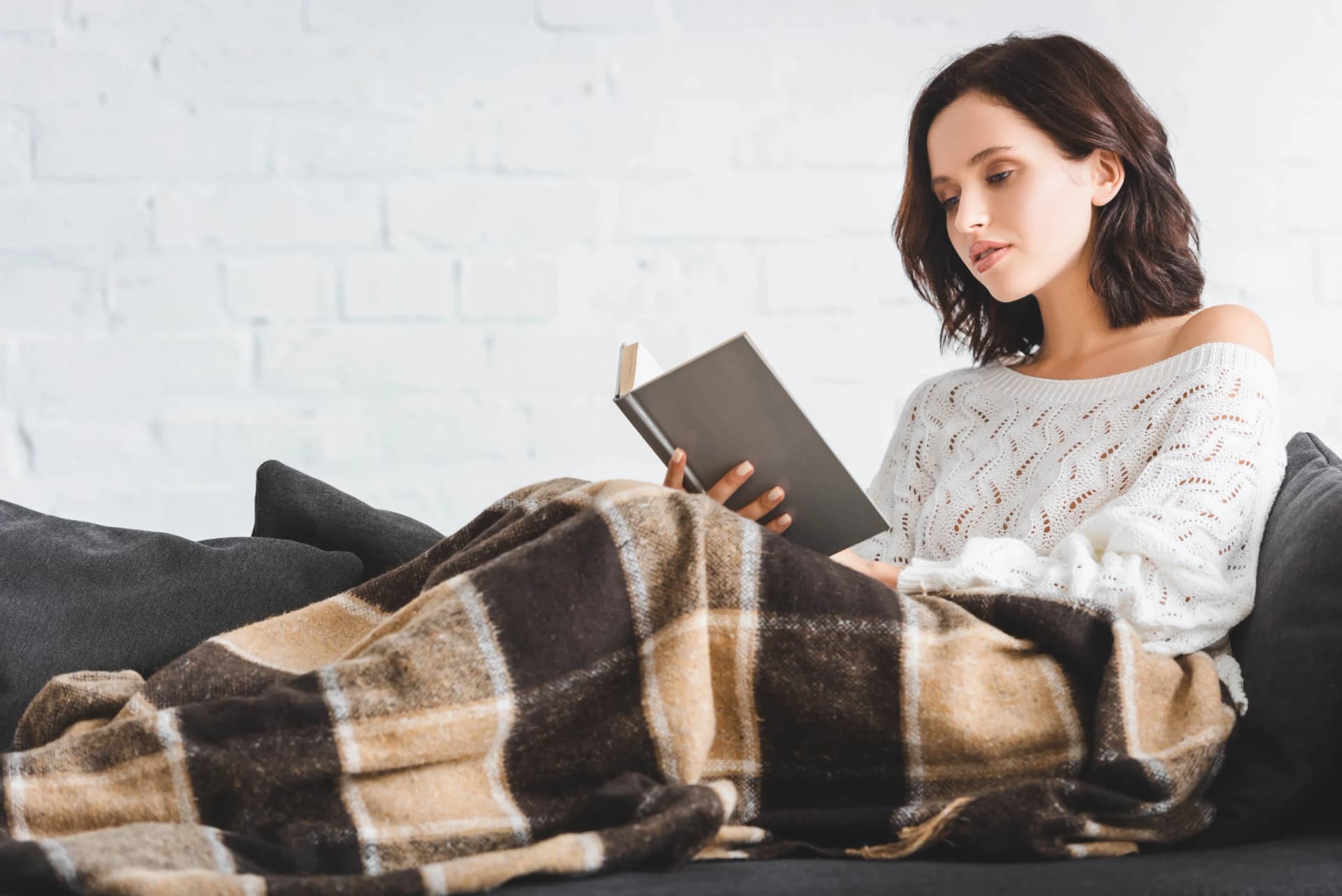 Young woman wrapped in a blanket reading a book on a sofa.