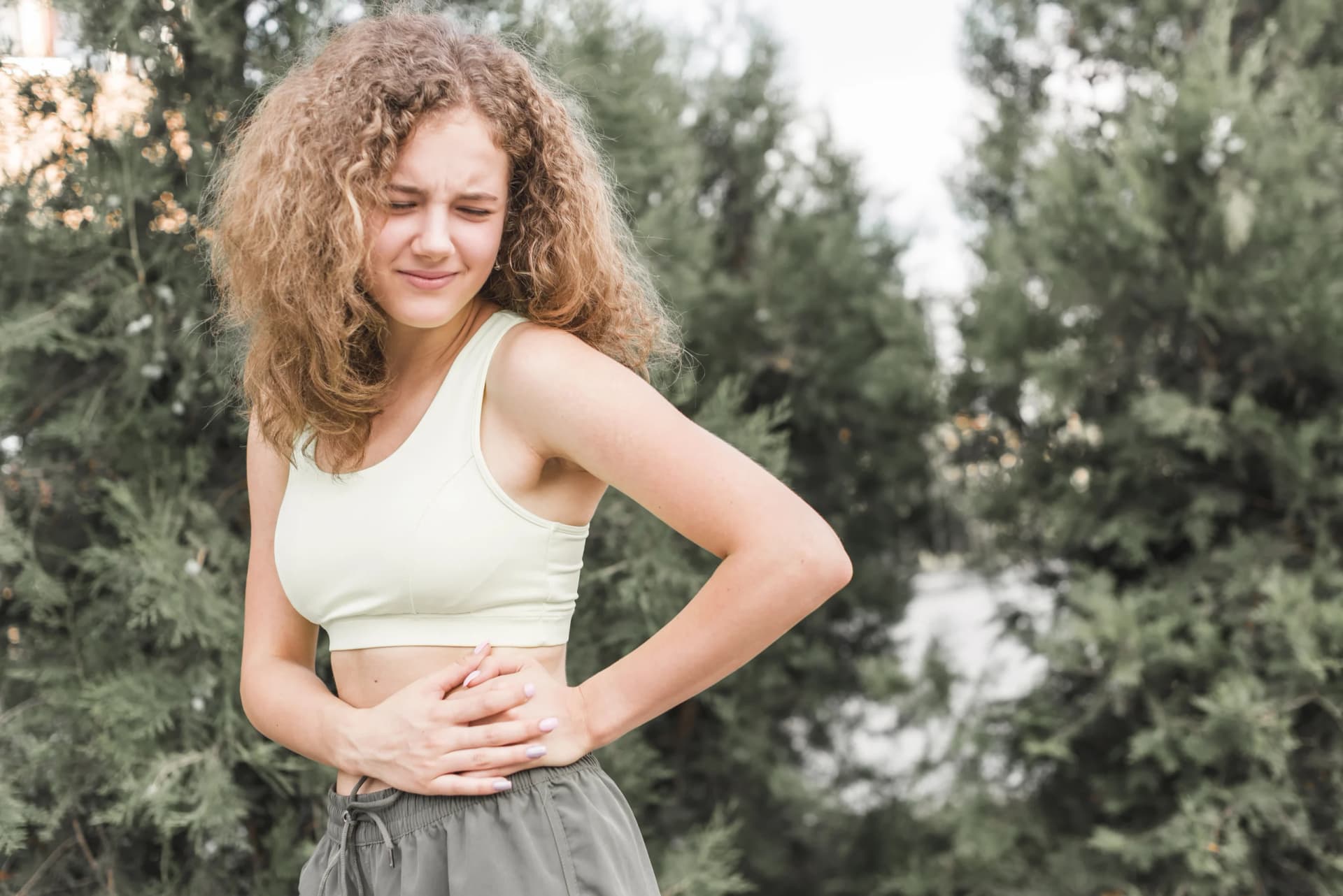 A young woman in activewear outdoors, holding her abdomen in discomfort.