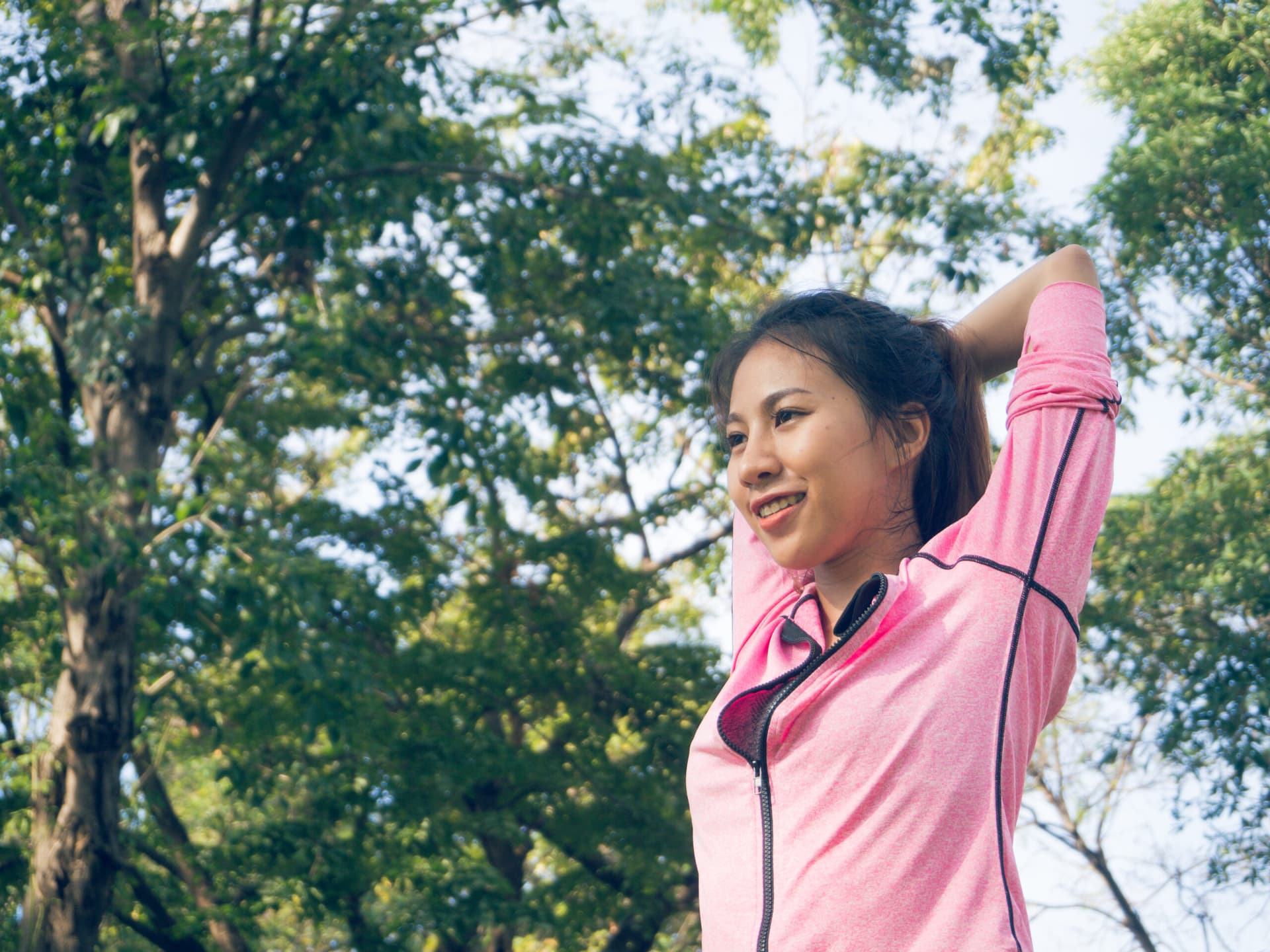 Woman in athletic wear stretching outdoors beneath tall trees.