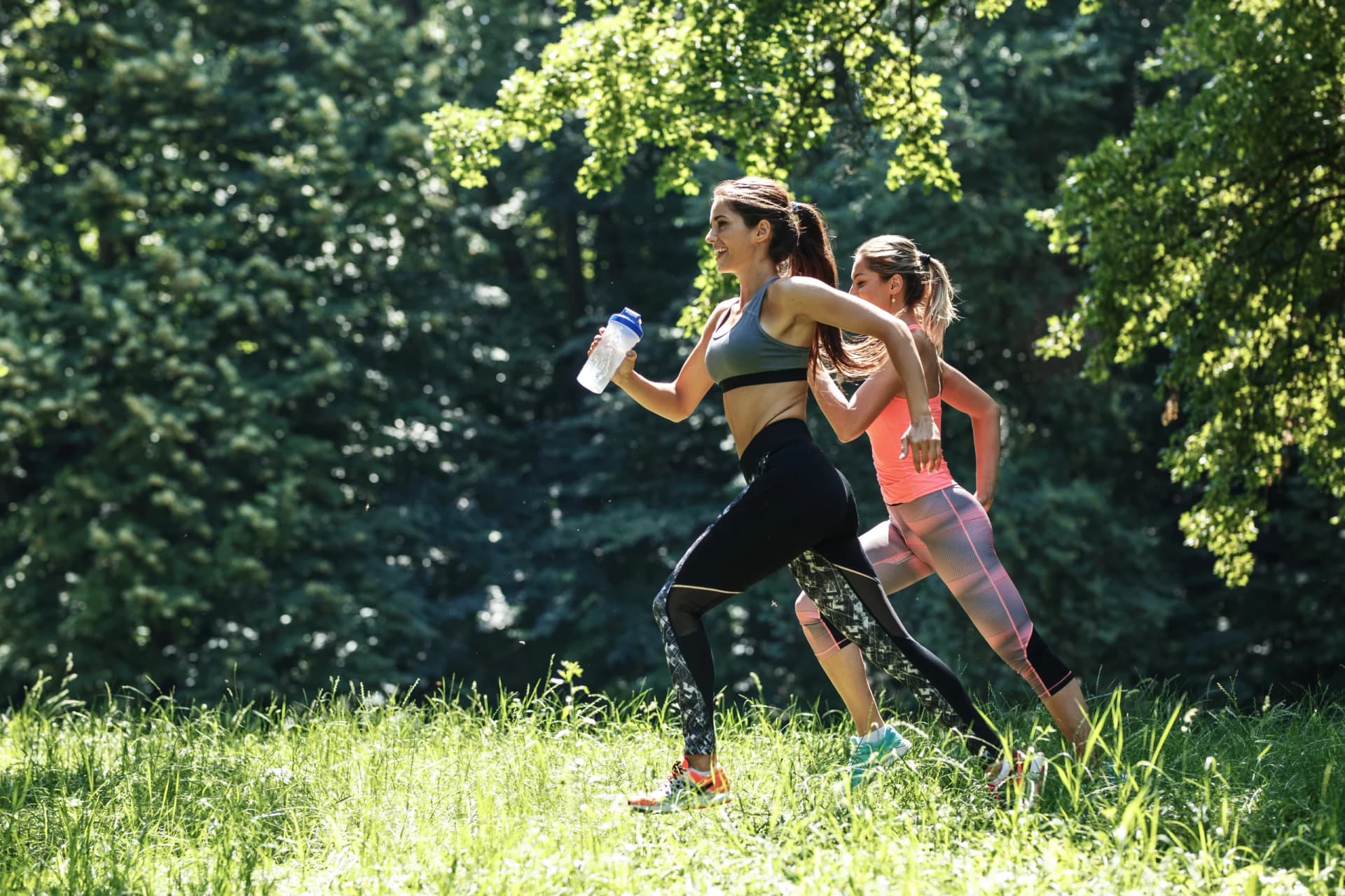 Two women jogging together along a sunlit forest trail.