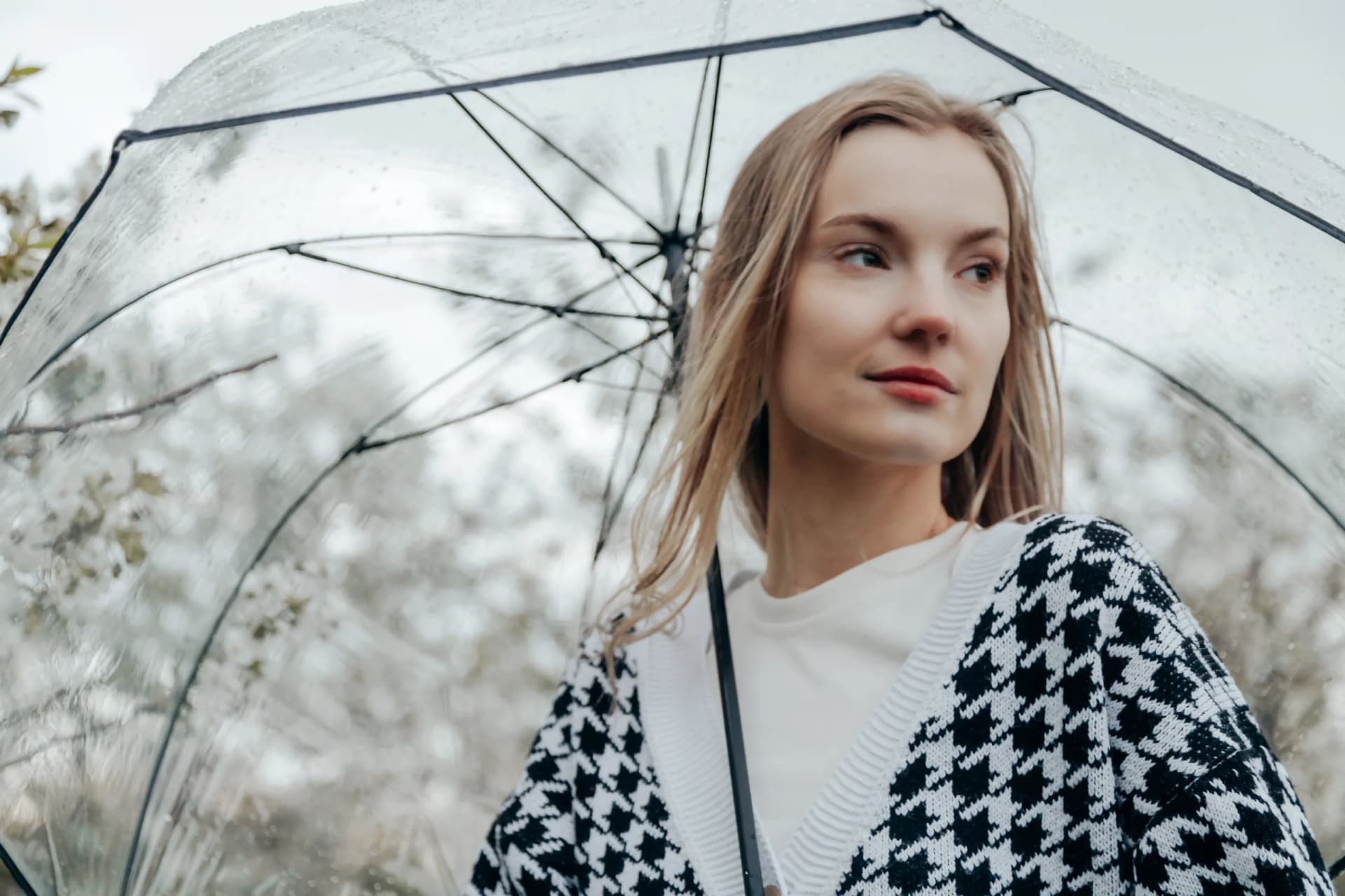 Woman in a plaid jacket standing under a transparent umbrella in the rain.