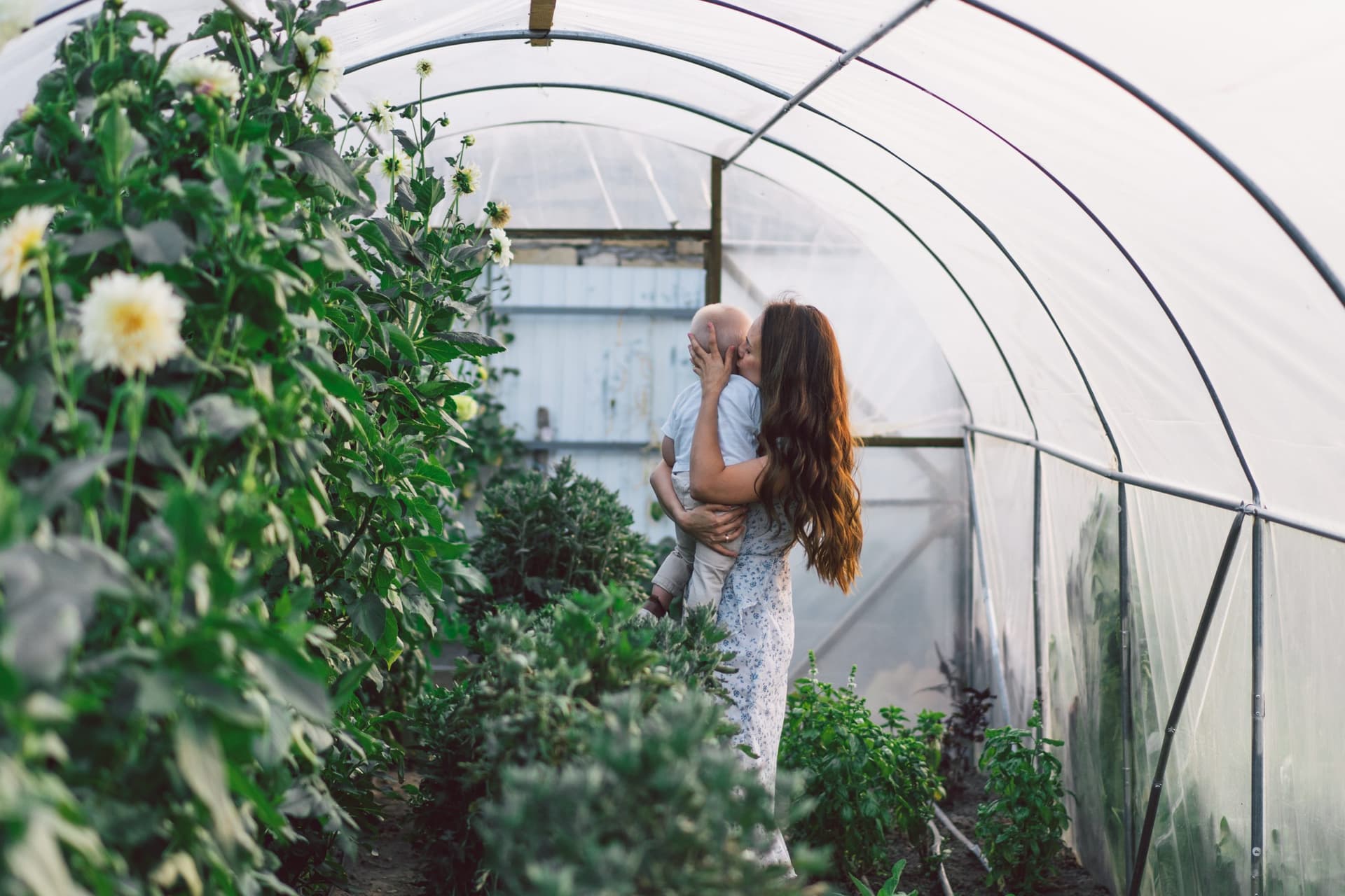 Mother kissing her young child while carrying them through a greenhouse full of flowers and herbs